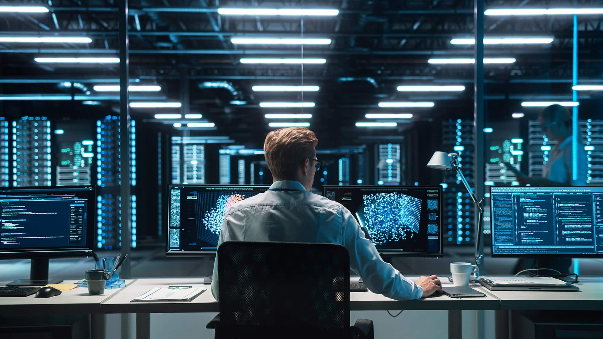 Software Developer Works with AI Analysis on his Computer with Two Monitors. Developer Coding while Looking at the Display of Computer. Modern Evening Dark Office. Back view.