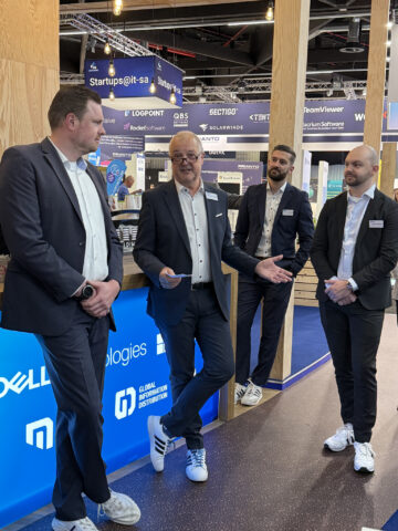 Vier Männer in Anzügen stehen an einem Stand einer Technologiemesse und unterhalten sich.