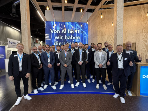 Eine Gruppe von Leuten in Anzügen und weißen Turnschuhen posiert auf einer Tech-Veranstaltung vor einem Display.