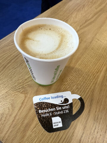 Eine Tasse Kaffee auf einem Holztisch neben einer Werbekarte in Form einer Kaffeetasse in deutscher Sprache.