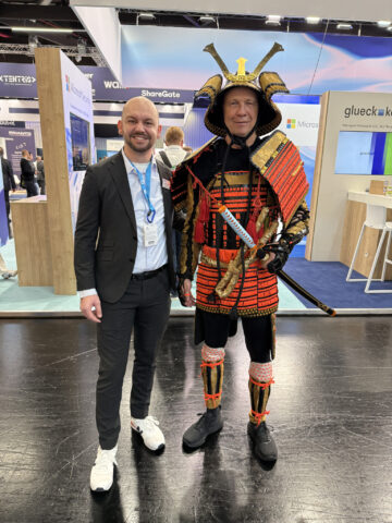 Zwei Männer posieren bei einer Veranstaltung; einer im Anzug, der andere in einer traditionellen Samurai-Rüstung.