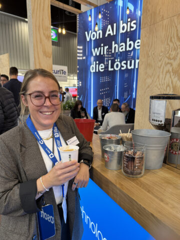 Frau hält eine Kaffeetasse und lächelt an einem Technikstand mit deutscher Beschilderung im Hintergrund.