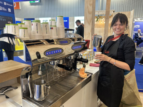 Ein Barista steht bei einer Veranstaltung an einer Kaffeemaschine und hält ein Milchkännchen und eine Tasse in der Hand.