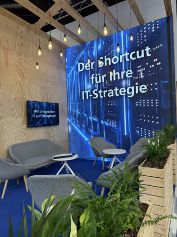 Moderner Lounge-Bereich mit grauen Stühlen, Pflanzen und einem großen Display, das für die IT-Strategie wirbt.