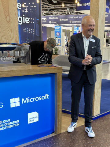 Zwei Männer stehen an einem Microsoft-Stand; einer lächelt im Anzug, der andere steht hinter einem Schalter.