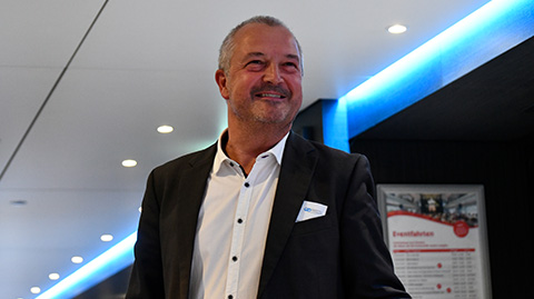 Ein Mann im Anzug steht in einem Innenraum unter blauer Akzentbeleuchtung und lächelt leicht.