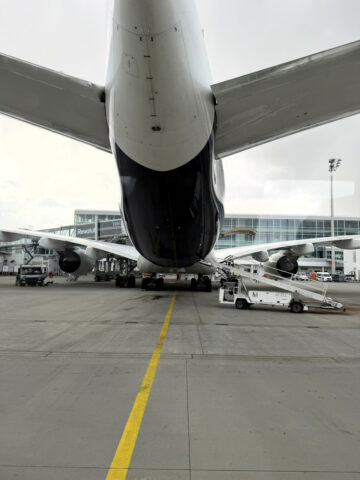 Rückansicht eines großen Flugzeugs, das an einem Flughafentor geparkt ist, mit Bodendienstfahrzeugen in der Nähe.