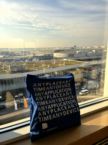 Eine blaue Tragetasche mit weißer Schrift liegt auf einer Fensterbank mit Blick auf ein Flughafenterminal.